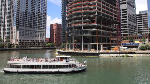 Workers install glass on a steel-frame skyscraper next to a river and a boat.